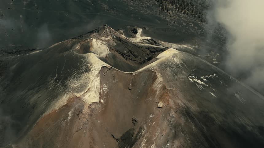 A dramatic, static aerial view looks down into the vast craters of a volcano as clouds drift through the barren landscape