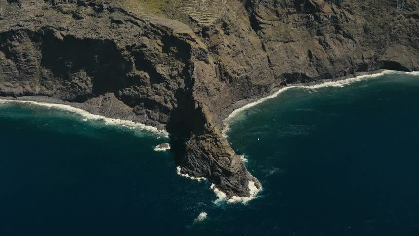 A static high-angle view of a dramatic sea stack jutting out from a rugged volcanic coastline into the deep blue ocean