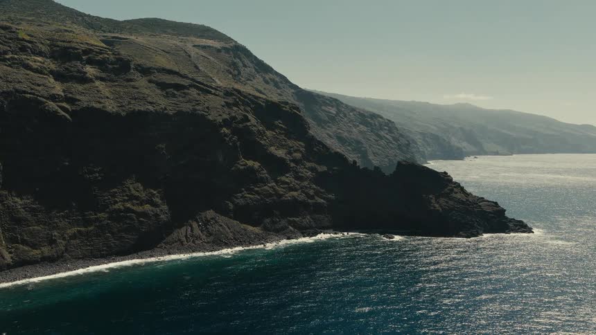 Serene wide shot of the dramatic, dark cliffs of a remote island coastline against the sparkling blue sea