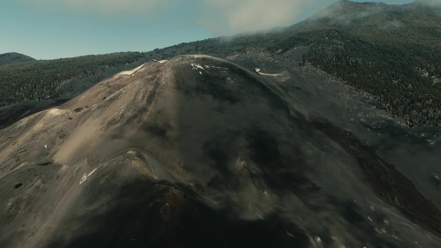 A wide, static view of a barren, ashen volcanic landscape, with a smoking, forest-covered mountain in the background