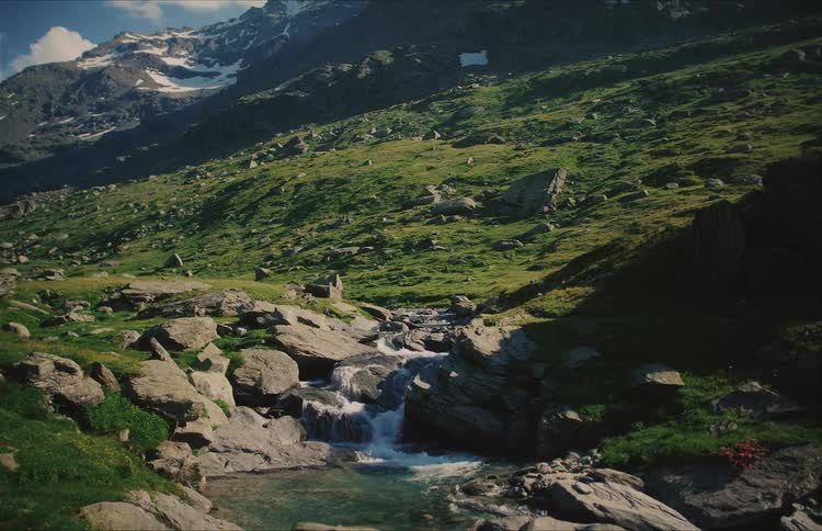 Peaceful wide shot of a small waterfall in a mountain river with lush green hills and snowy mountains in the background