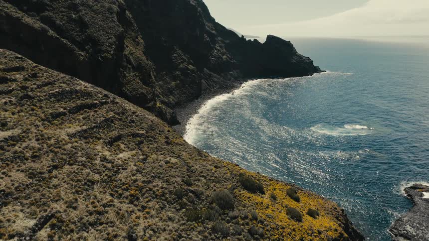 A slow aerial ascent reveals a vast, rugged volcanic coastline with sparse yellow vegetation meeting the deep blue ocean