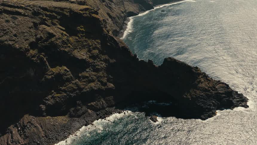 A dramatic high-angle view of a rugged, volcanic coastline where powerful ocean waves crash against the dark, rocky cliffs