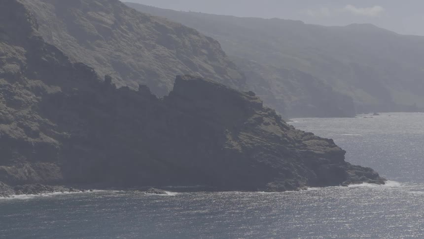 A slow zoom-out reveals the vast and rugged coastline of a misty volcanic island meeting the sparkling sea