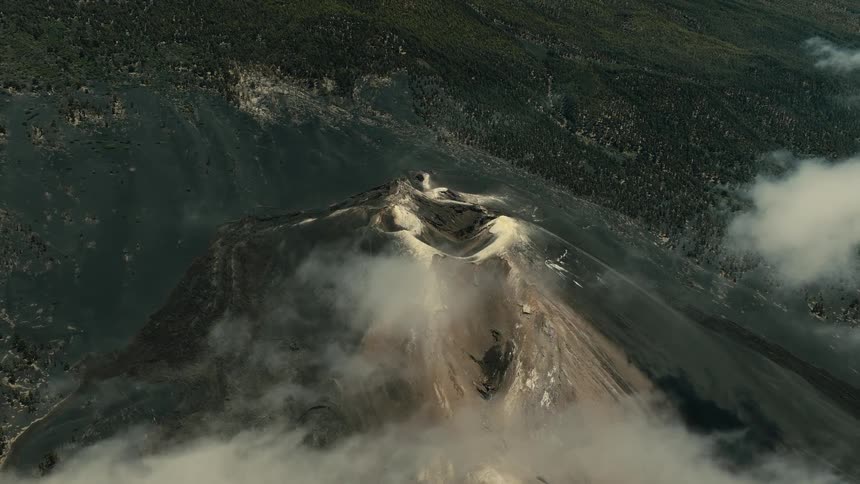 An epic high-angle aerial shot reveals a massive volcano crater with wisps of clouds drifting over its barren, ashen slopes and surrounding forest