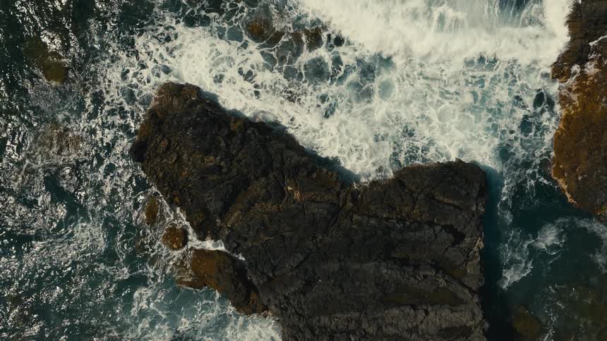 A powerful ocean wave crashes against a dark volcanic rock formation in dramatic slow motion, as seen from a top-down aerial perspective