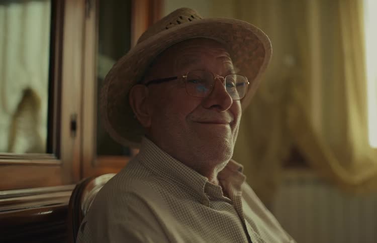 A contented elderly man in a straw hat and glasses smiles softly while sitting indoors in the warm afternoon light