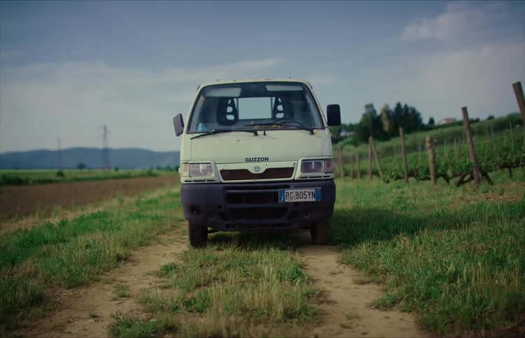 An old white van is parked on a dirt road next to a vineyard in the rustic Italian countryside on an overcast day