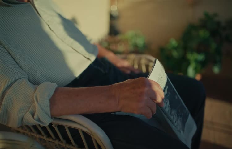 A close-up of a senior man's hands as he reads a newspaper while relaxing in a sunlit chair
