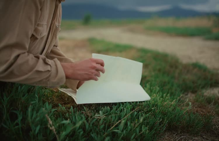 An explorer kneels in a grassy field to study a large map before carefully folding it up