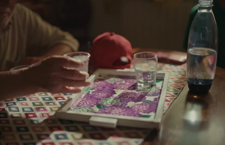 An older man sitting at a table with a patterned tablecloth slowly reaches for and picks up a plastic cup of water
