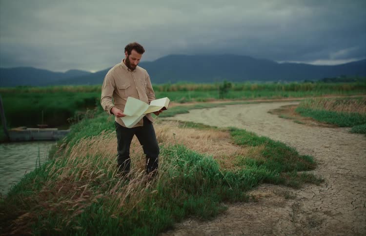 A lone bearded traveler unfolds a hidden map from a book while standing on a grassy path in a remote valley under a cloudy sky