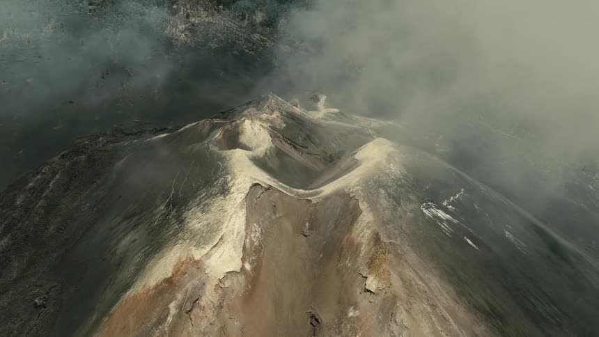 A dramatic high-angle aerial view shows a massive volcanic crater being slowly covered by thick, rolling clouds