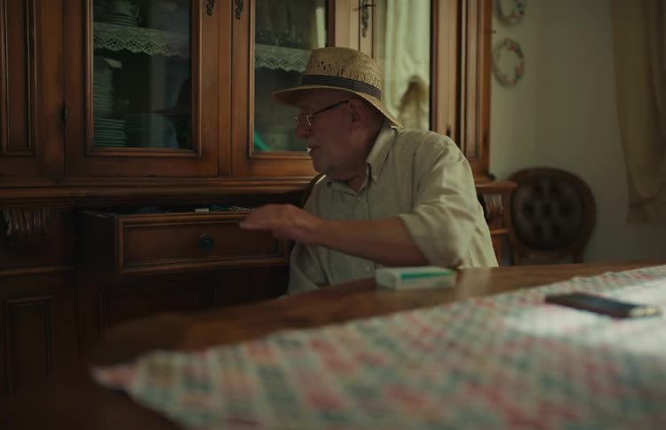 Elderly man wearing a hat and glasses sits at a dining table and searches for something in a cabinet drawer