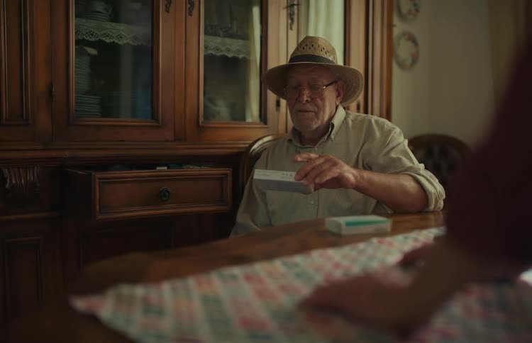 A senior man wearing a straw hat pulls his medication from a wooden cabinet drawer at the dining table