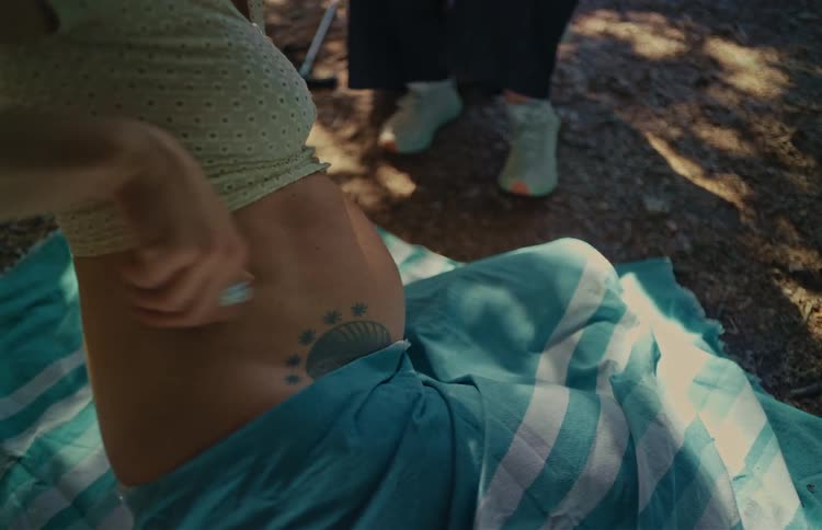 A pregnant woman reveals a sun tattoo as she rubs her belly while sitting on a blanket outside