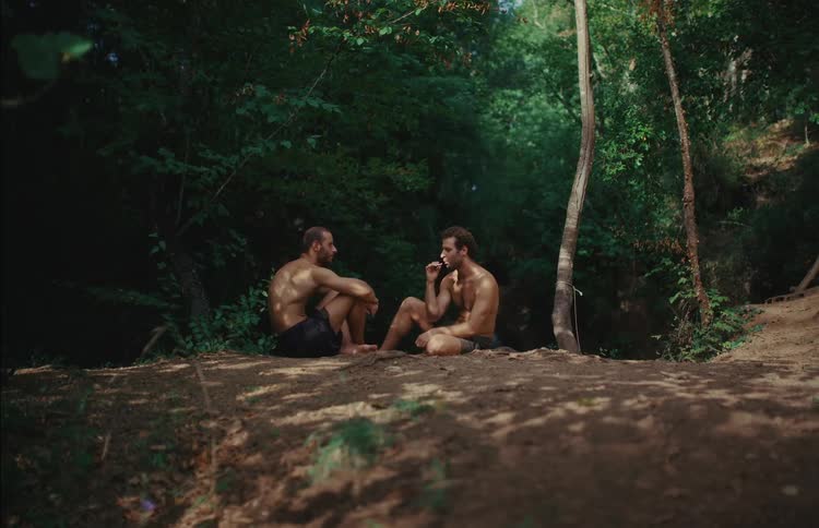 Two shirtless men sit on a dirt path in a serene forest, talking and sharing a moment of camaraderie with a friendly handshake