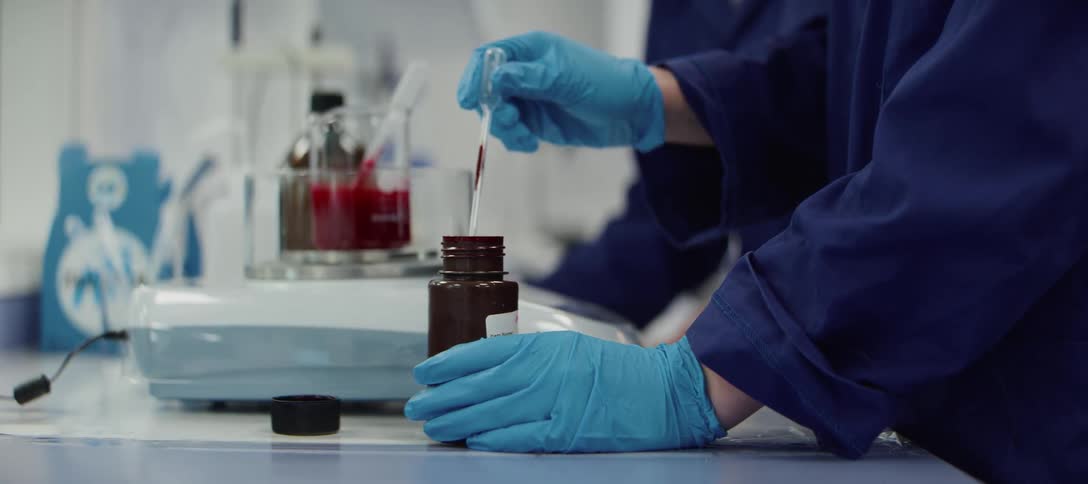 A scientist in a lab carefully transfers red liquid from a bottle into a beaker using a pipette in slow motion