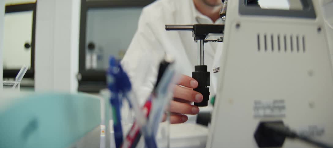 A scientist's hand is seen in close-up as they carefully adjust the focus knob of a microscope in a research lab