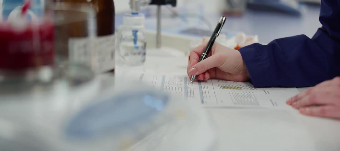 A scientist in a blue lab coat meticulously records data on a chart in a sterile laboratory setting