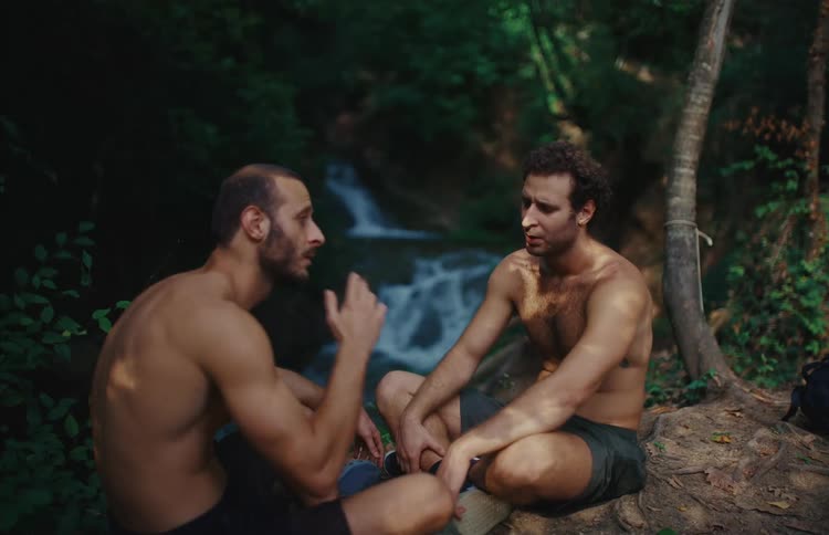 Two shirtless male hikers sit and talk by a waterfall in a lush forest, with one smoking a cigarette