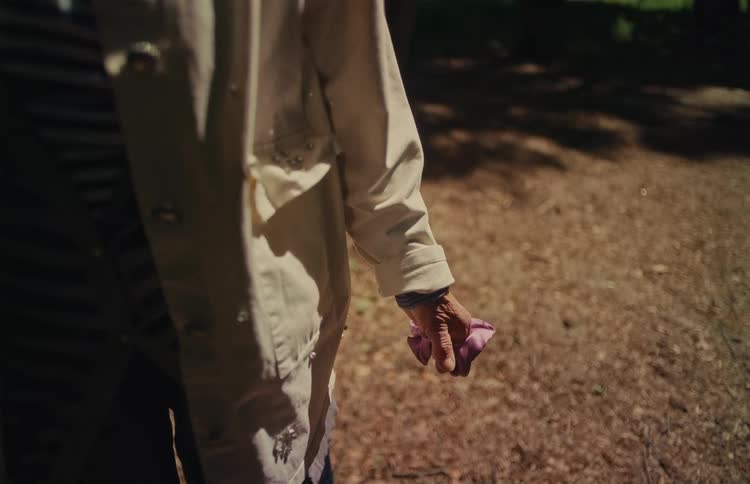 Static close-up shot of an elderly person's hand clutching a pink cloth while standing in a sun-dappled forest