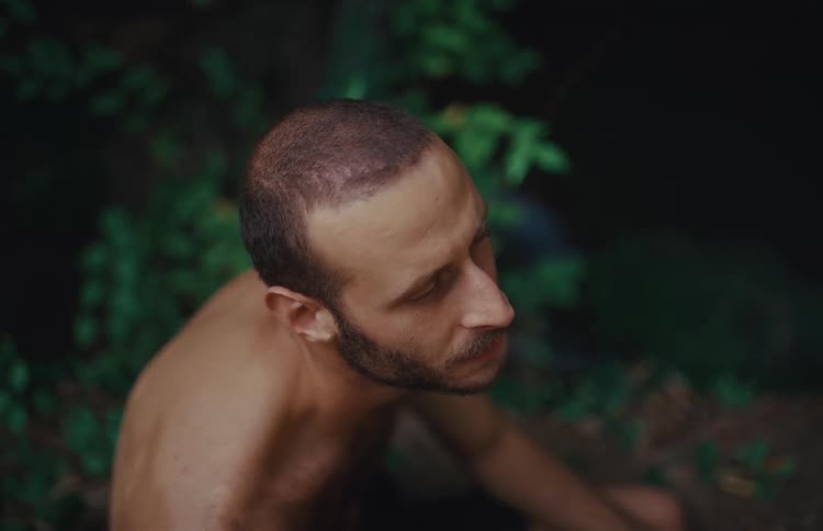 A shirtless man sits alone in the woods, smoking and looking around with a thoughtful and dazed expression in slow motion