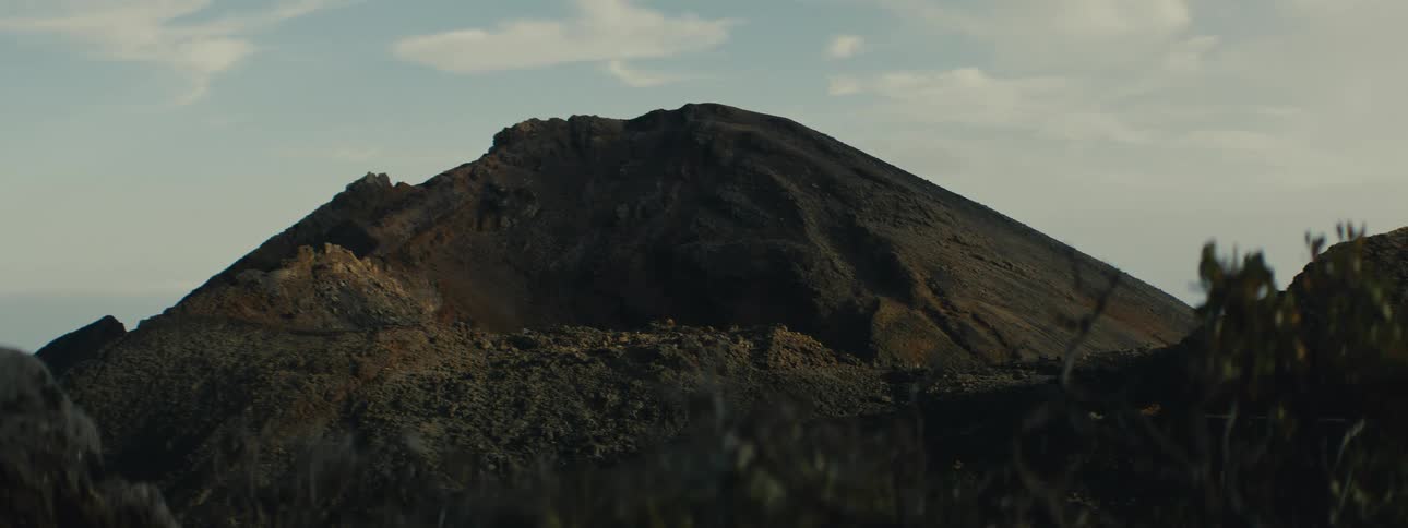 A cinematic close-up of a dark volcanic mountain peak under a soft, hazy sky
