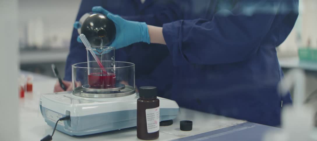 A scientist in a blue lab coat carefully pours a red liquid from a flask into a beaker on a precision scale