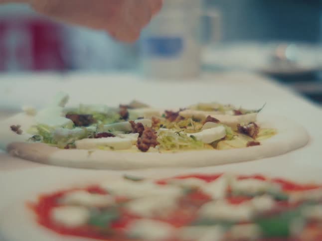 A chef's hands carefully place fresh greens, cheese, and walnuts onto an uncooked white pizza base