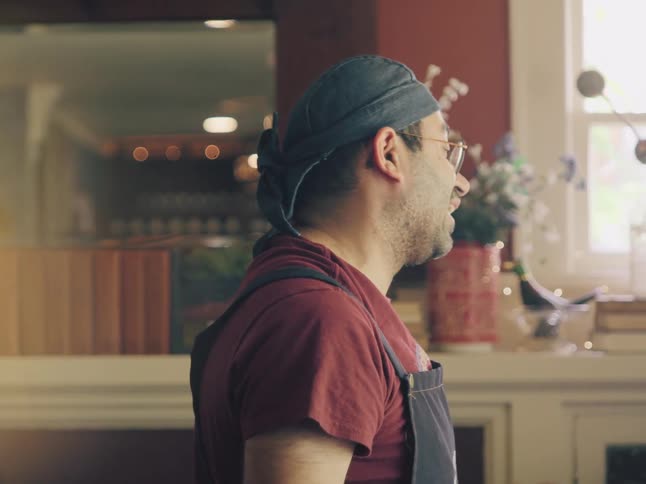 A profile shot of a man wearing a bandana and apron while talking to someone off-screen in a warmly lit interior
