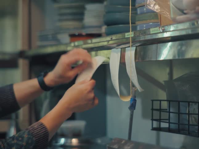 A server's hands hang a new order ticket on a stainless steel rack in a restaurant kitchen