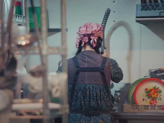 A restaurant worker wearing a bandana and apron washes colorful trays in a commercial kitchen sink