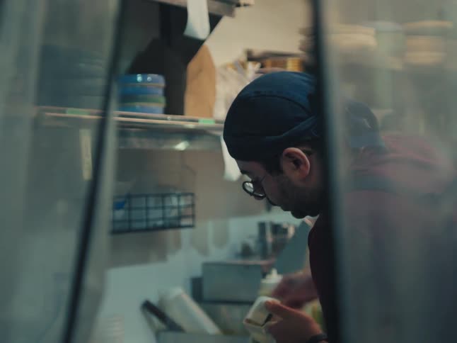A chef carefully prepares a food order in a commercial kitchen, viewed from behind a plastic curtain