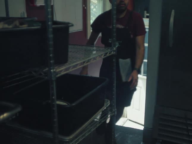 A chef in an apron walks up the stairs from a bright doorway into a dark kitchen storage room
