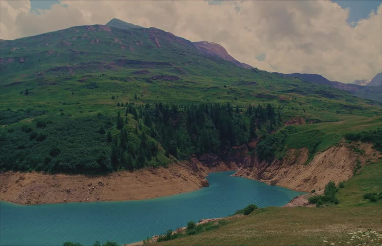 A picturesque, static wide shot of a vibrant turquoise alpine lake nestled within lush green mountains under a partly cloudy sky