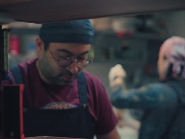 Two chefs in aprons and bandanas work together preparing food in a bustling, dimly lit restaurant kitchen