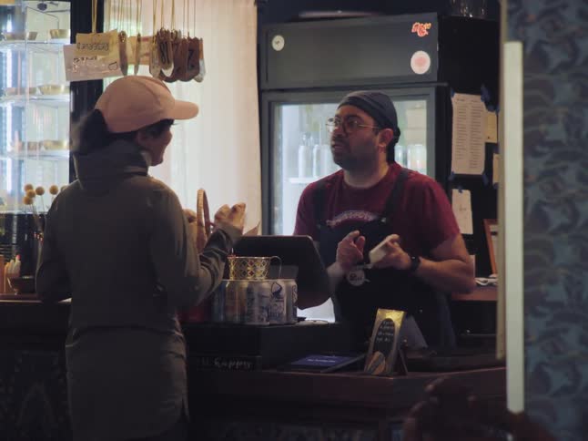 A woman in a hat smiles as she orders from a friendly barista at a small, cozy cafe counter
