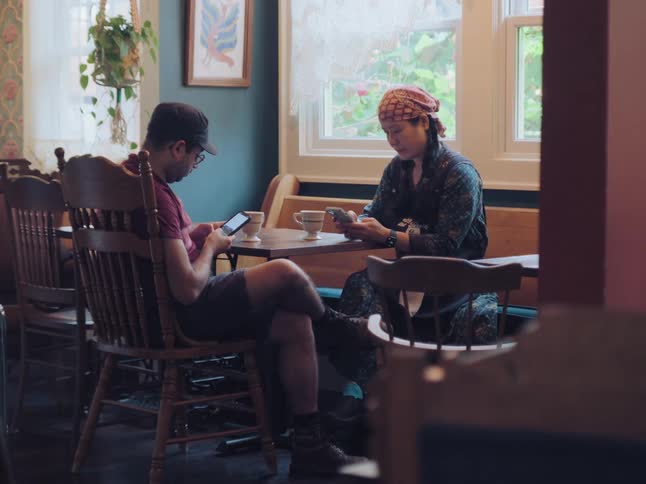 A man and a woman sit in silence at a coffee shop, both absorbed in their smartphones and ignoring each other