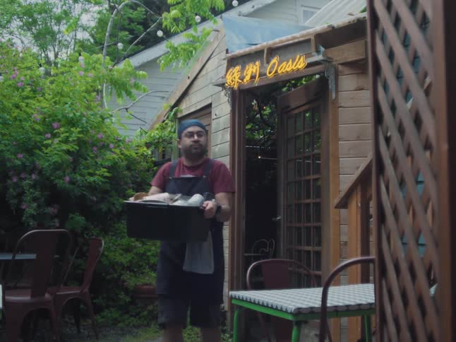 A restaurant worker carries a bin of dirty dishes in slow motion across a lush garden patio with a neon sign