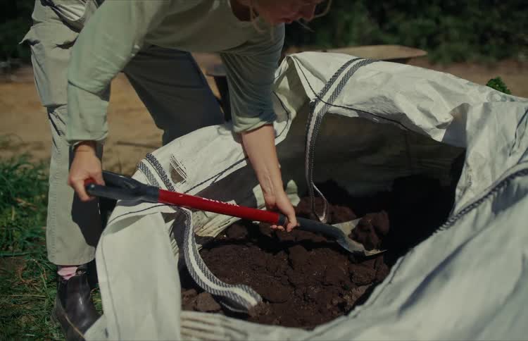 A gardener shovels dark, rich compost out of a large white bag outdoors on a sunny day