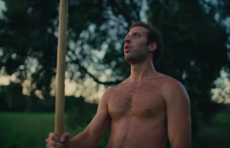 A muscular, shirtless man takes a deep breath after an intense sledgehammer workout in a field at dusk