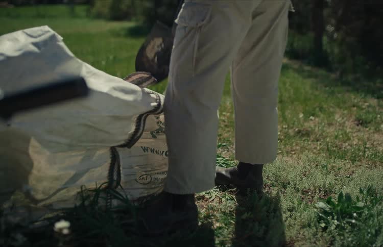 A person in work boots shovels soil from a large bag onto a grassy yard in slow motion