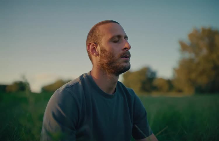 A pensive man sits in a grassy field and closes his eyes to meditate at dusk