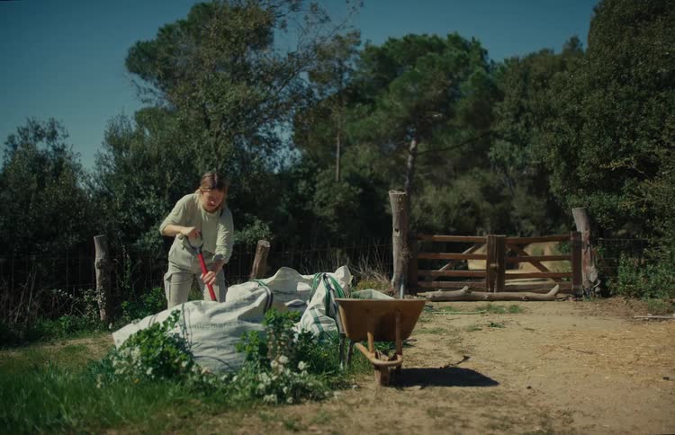 A woman shovels compost or soil from a large sack into a wheelbarrow on a sunny day in the countryside