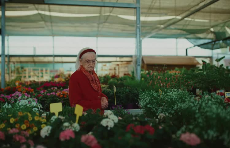 A thoughtful elderly woman in a red cardigan admires the variety of colorful potted flowers in a plant nursery