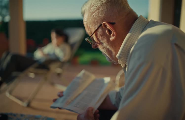 Elderly man with glasses finds a moment of peace reading a book on the patio while his wife relaxes in the background