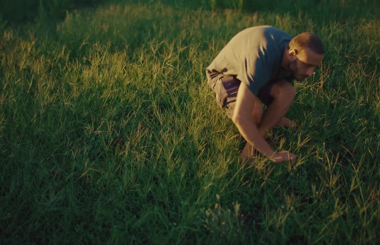 A barefoot man walks through a sunlit field and sits down to contemplate on the lush green grass