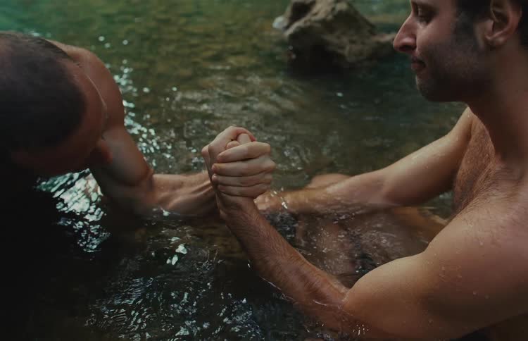 Two shirtless men have an intense arm wrestling match while sitting in the cool water of a river