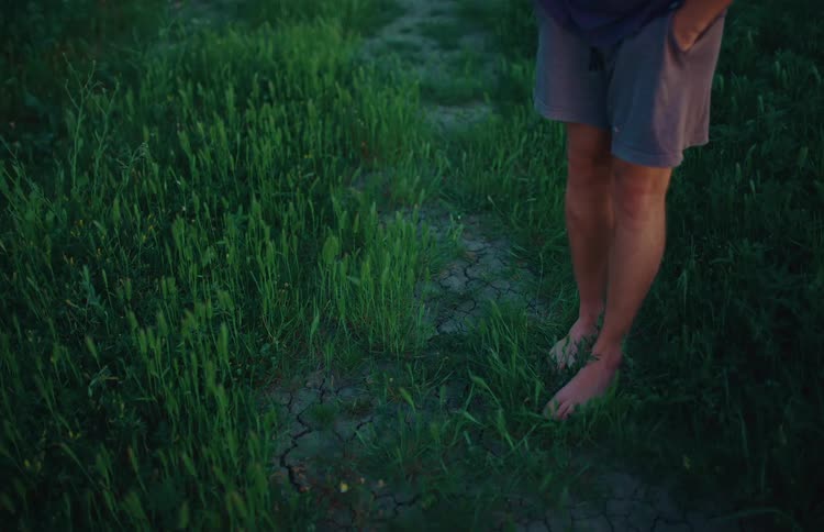 A man stands barefoot with his hands in his pockets in a grassy field at dusk